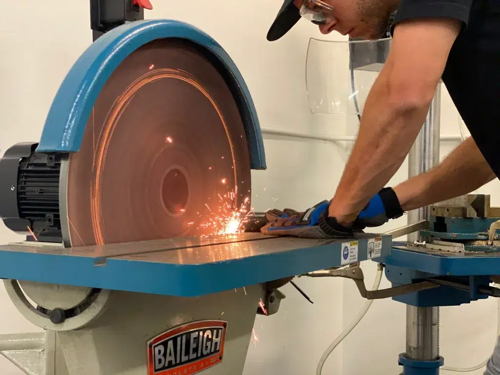 A person wearing gloves and safety glasses grinds metal on a Baileigh industrial metalworking machine by JPW Industries, creating sparks.