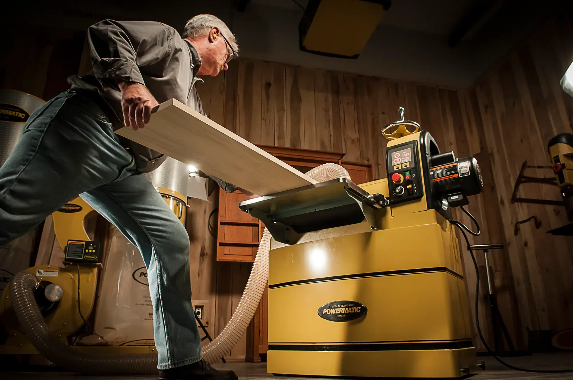 A man feeds a wooden board into a yellow Powermatic industrial woodworking machine by JPW Industries inside a workshop with wood-paneled walls.